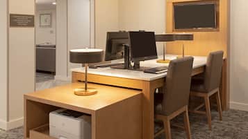 A hotel room with a desk, two chairs, a flat-screen TV mounted on the wall, a desk lamp, and a printer.