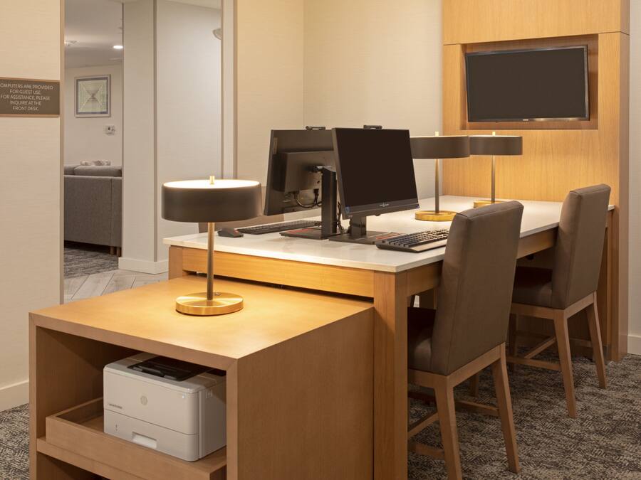 A hotel room with a desk, two chairs, a flat-screen TV mounted on the wall, a desk lamp, and a printer.