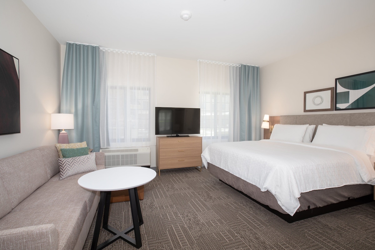 A hotel room with a bed, a sofa, a TV, a small table, and a window with curtains.
