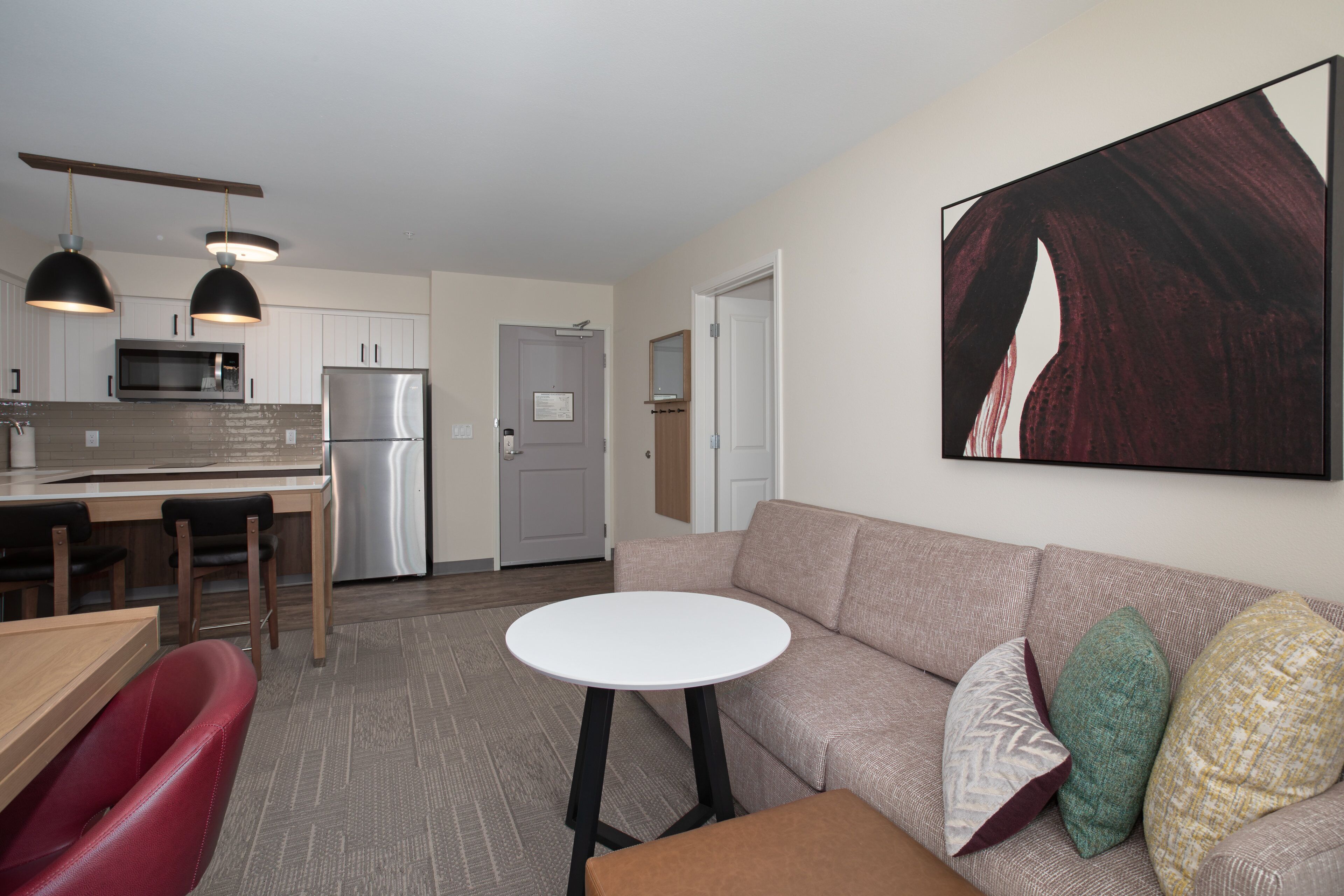 A modern living room with a sofa, a small round table, and a kitchen area with a refrigerator, microwave, and wooden cabinets.