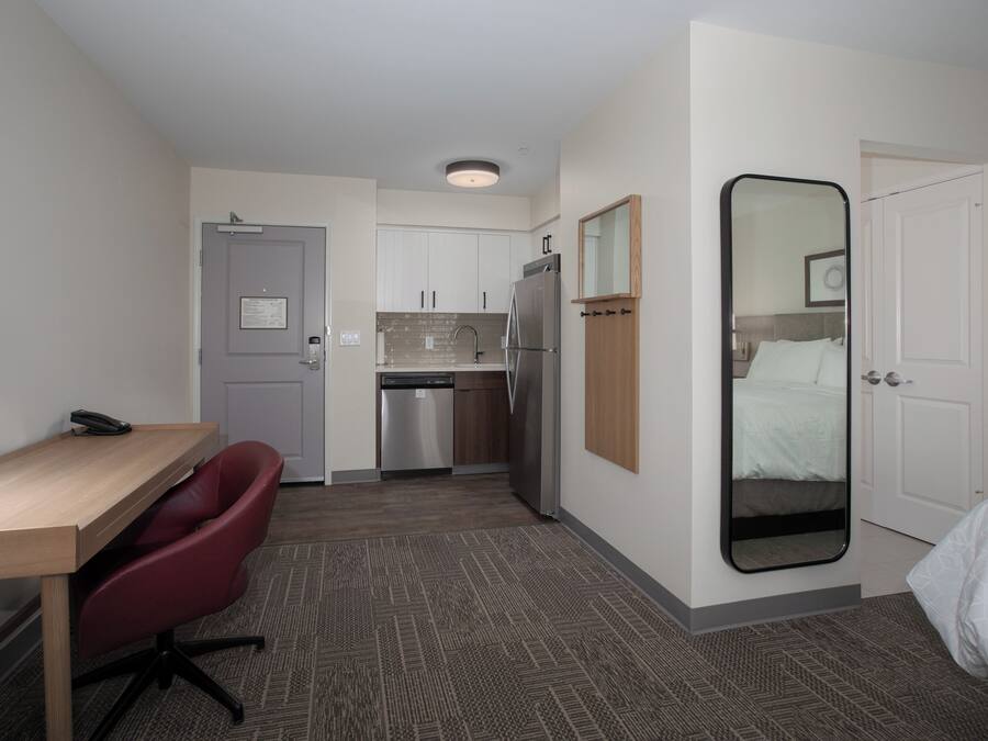 A compact hotel room with a kitchenette, a desk with a chair, a bed, and a large mirror.