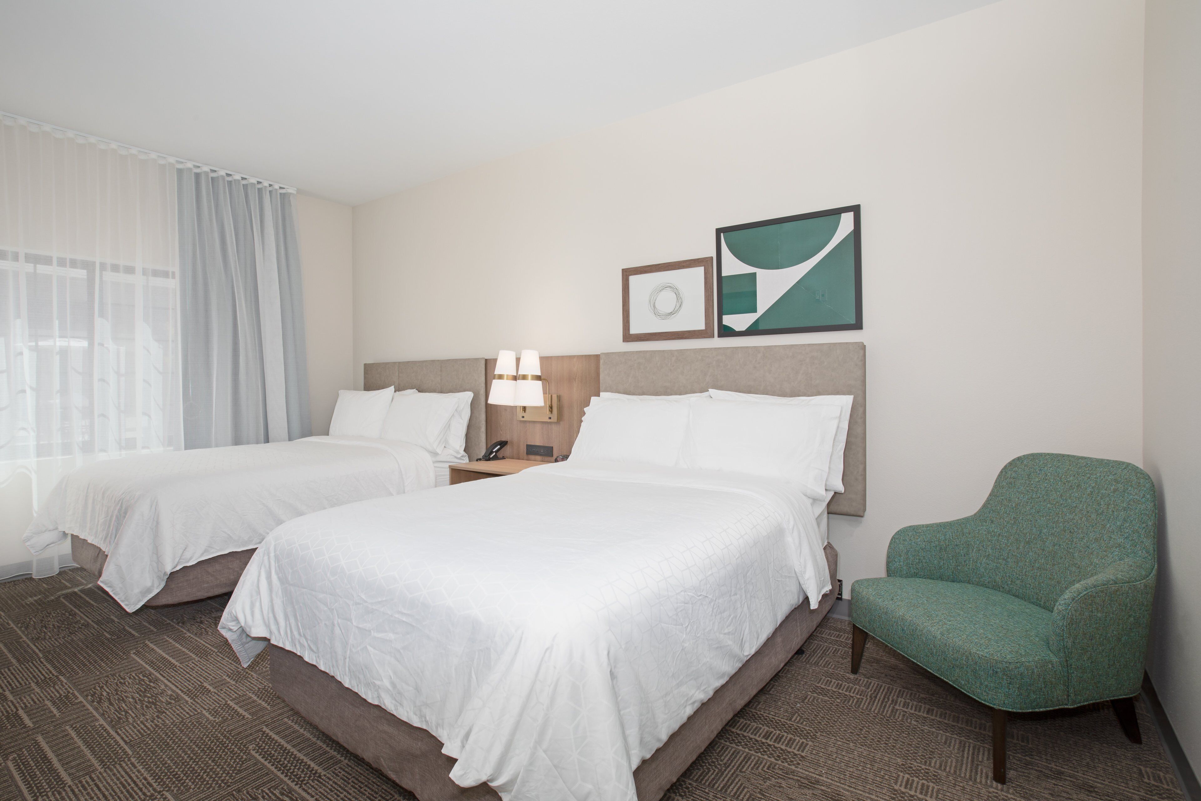 A hotel room with two beds, a green armchair, a nightstand with a lamp, and a framed picture on the wall.