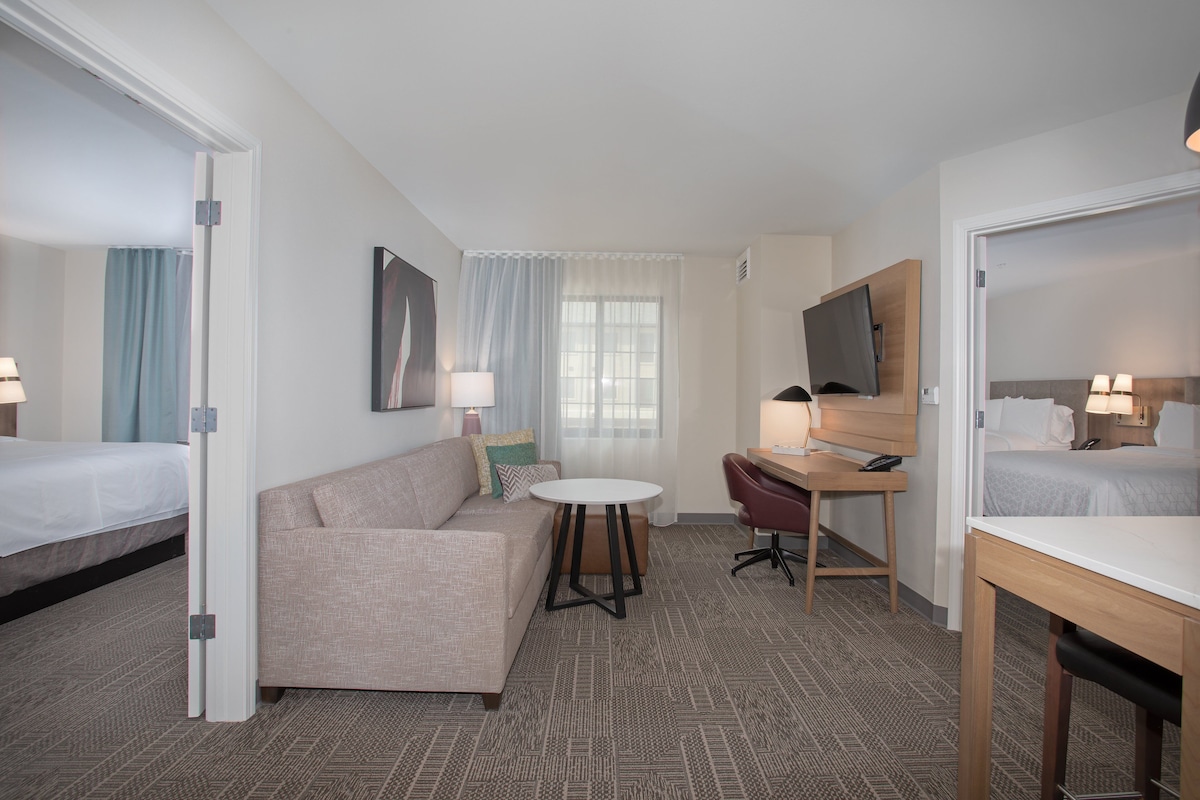 A hotel room with a bed, a sofa, a desk, a chair, a small table, a mirror, and a window with curtains.