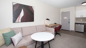 Una sala de estar con sofá, una mesa redonda, una silla roja, un escritorio y una zona de cocina con armarios y un refrigerador.