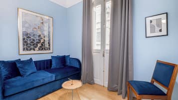 A room with a blue sofa, a wooden chair, a small round table, and two framed artworks on the wall.