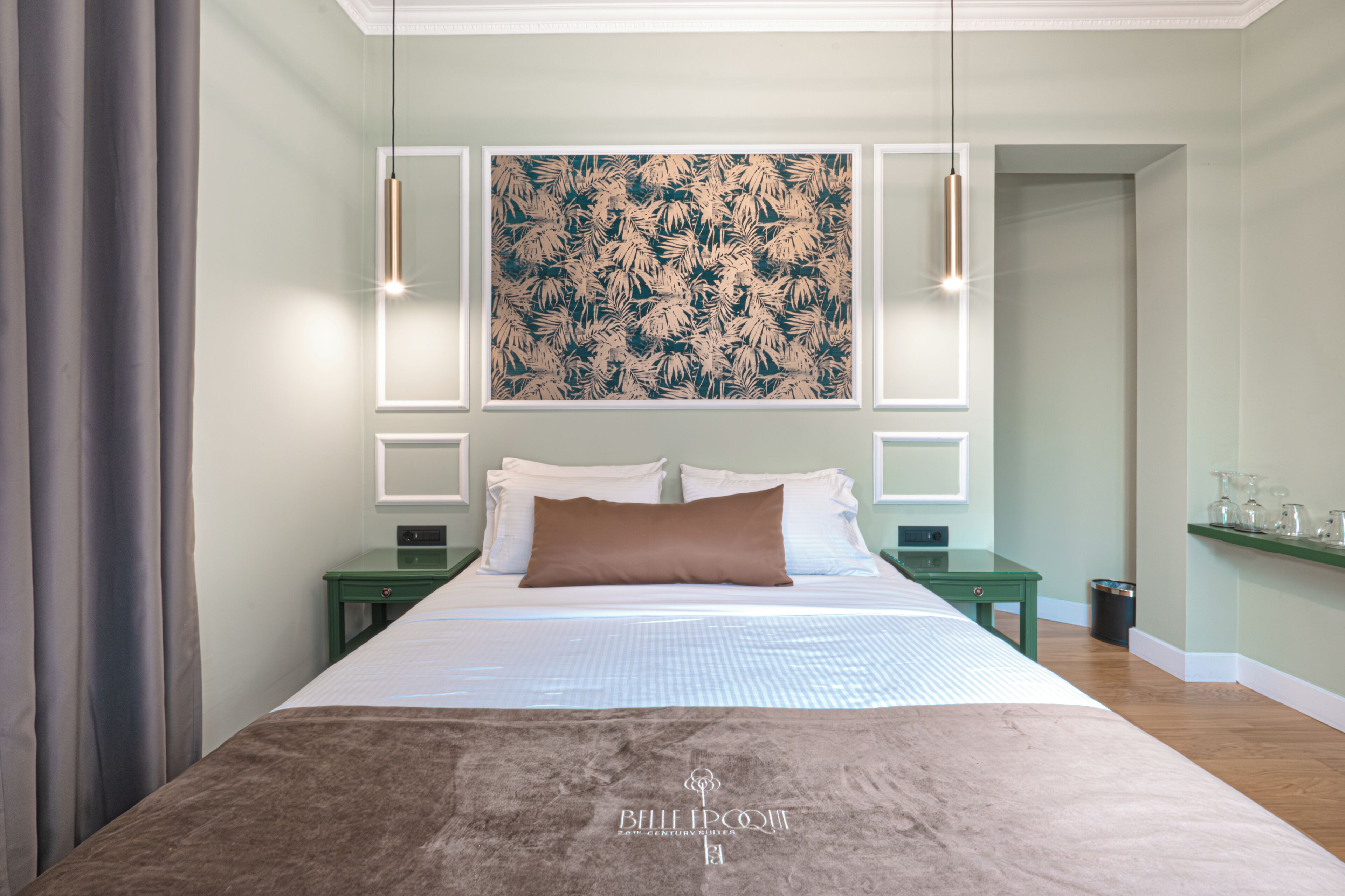 A hotel room with a large bed, a nightstand, a green side table, a wall-mounted artwork, and a hanging light fixture.