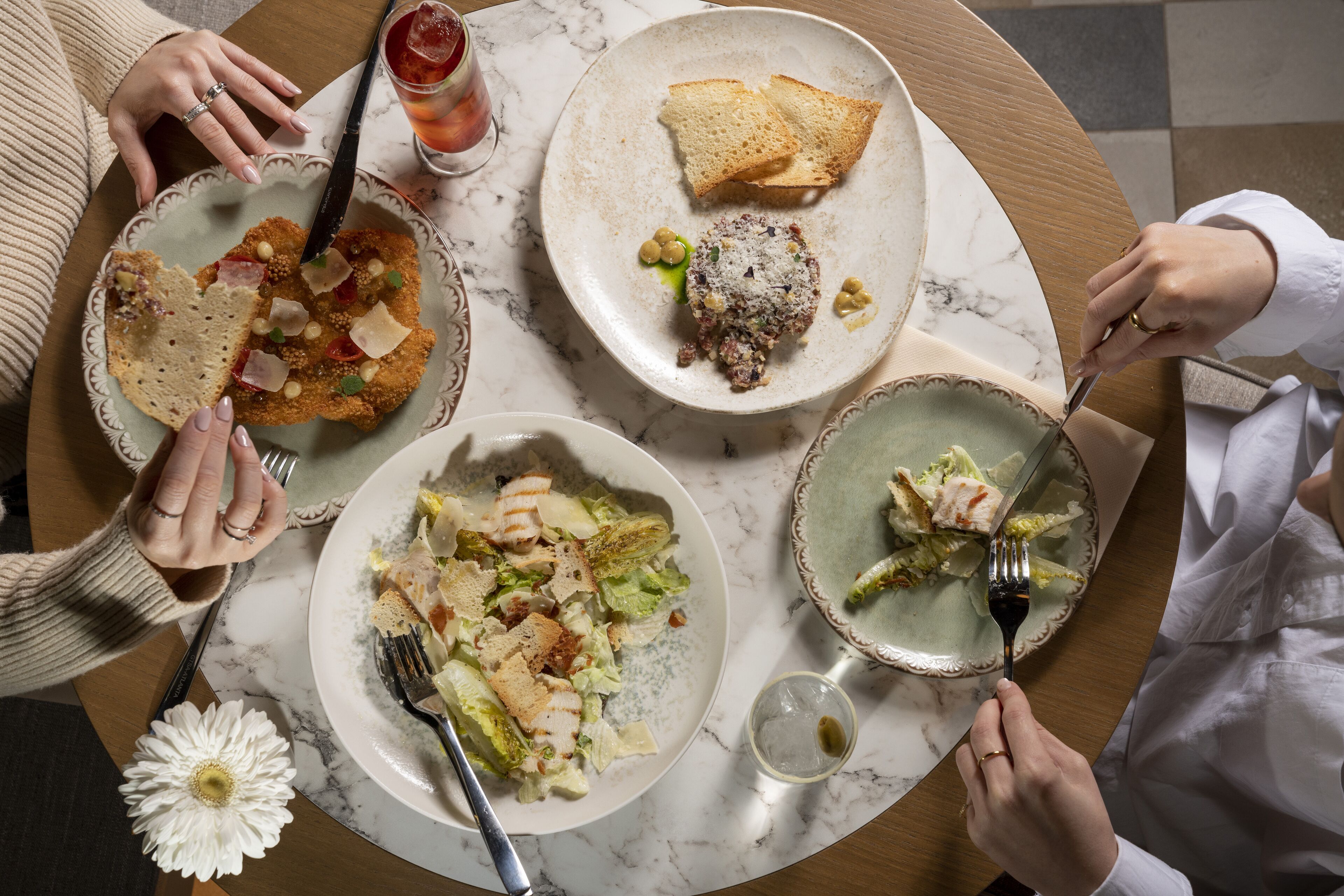 Een tafel met verschillende gerechten, zoals brood, salade en een vleesgerecht, omringd door mensen.