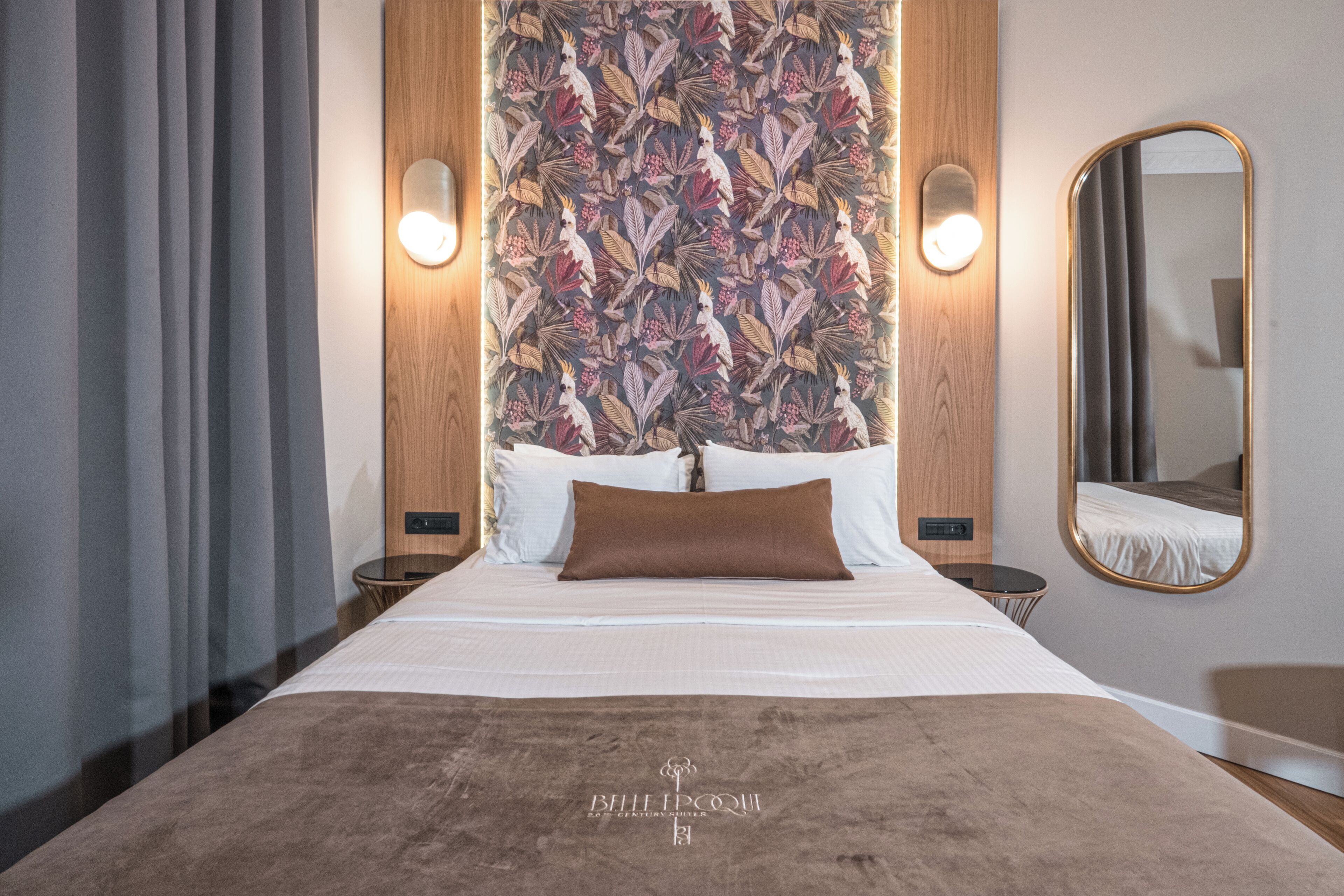 A hotel room with a large bed, a headboard with a floral pattern, two bedside lamps, a round mirror, and a wooden accent wall.