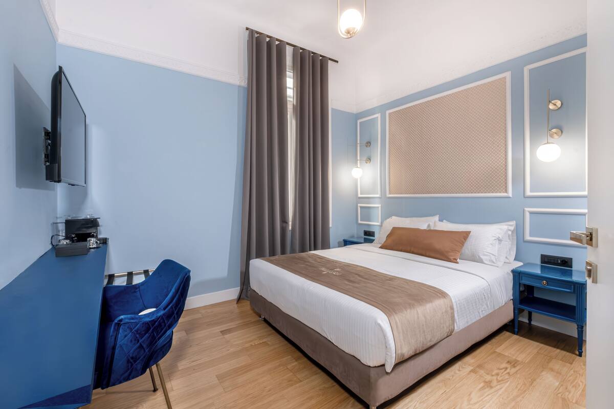 A modern hotel room with a large bed, a desk with a chair, a TV mounted on the wall, and a window with curtains.