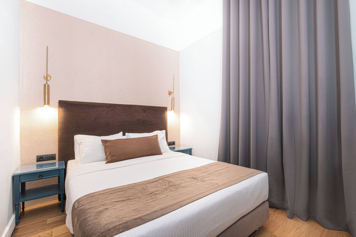 A neatly made bed with a brown headboard, two bedside tables with lamps, and a large window with curtains.