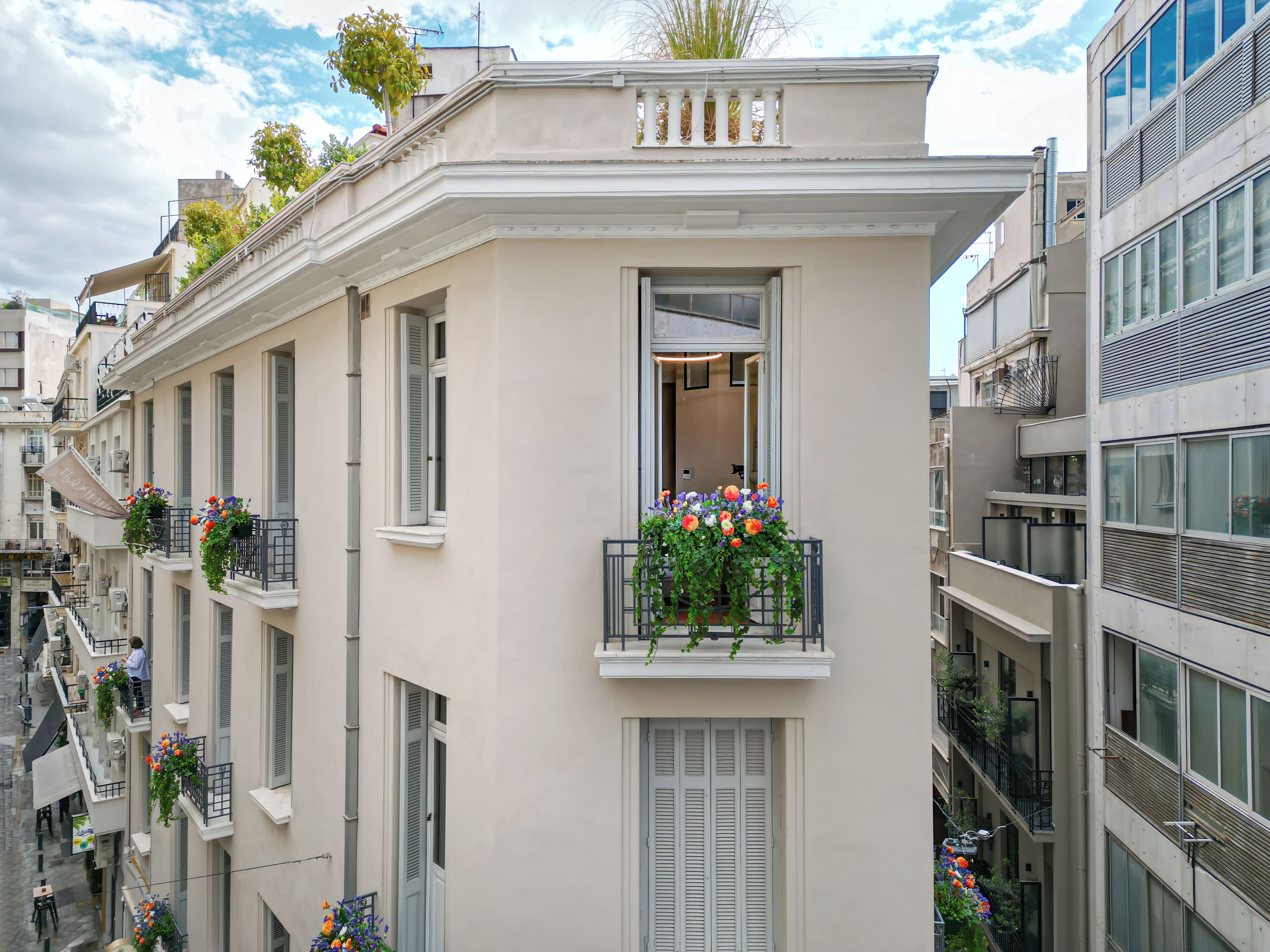 A multi-story building with balconies adorned with flowers, a central entrance, and a view of adjacent buildings.