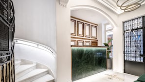 Um hall de hotel com uma escadaria de mármore, uma receção com frente em mármore verde e um portão de metal decorativo.