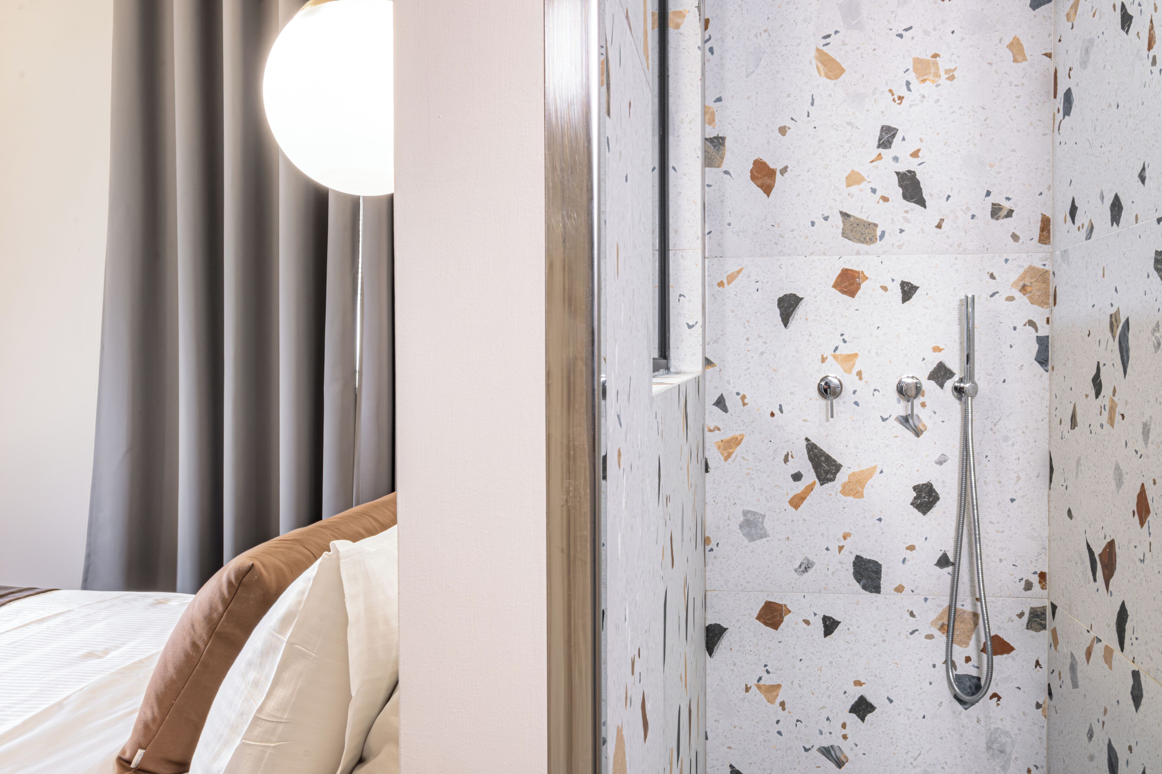 A hotel room with a bed, curtains, and a shower area with terrazzo flooring.