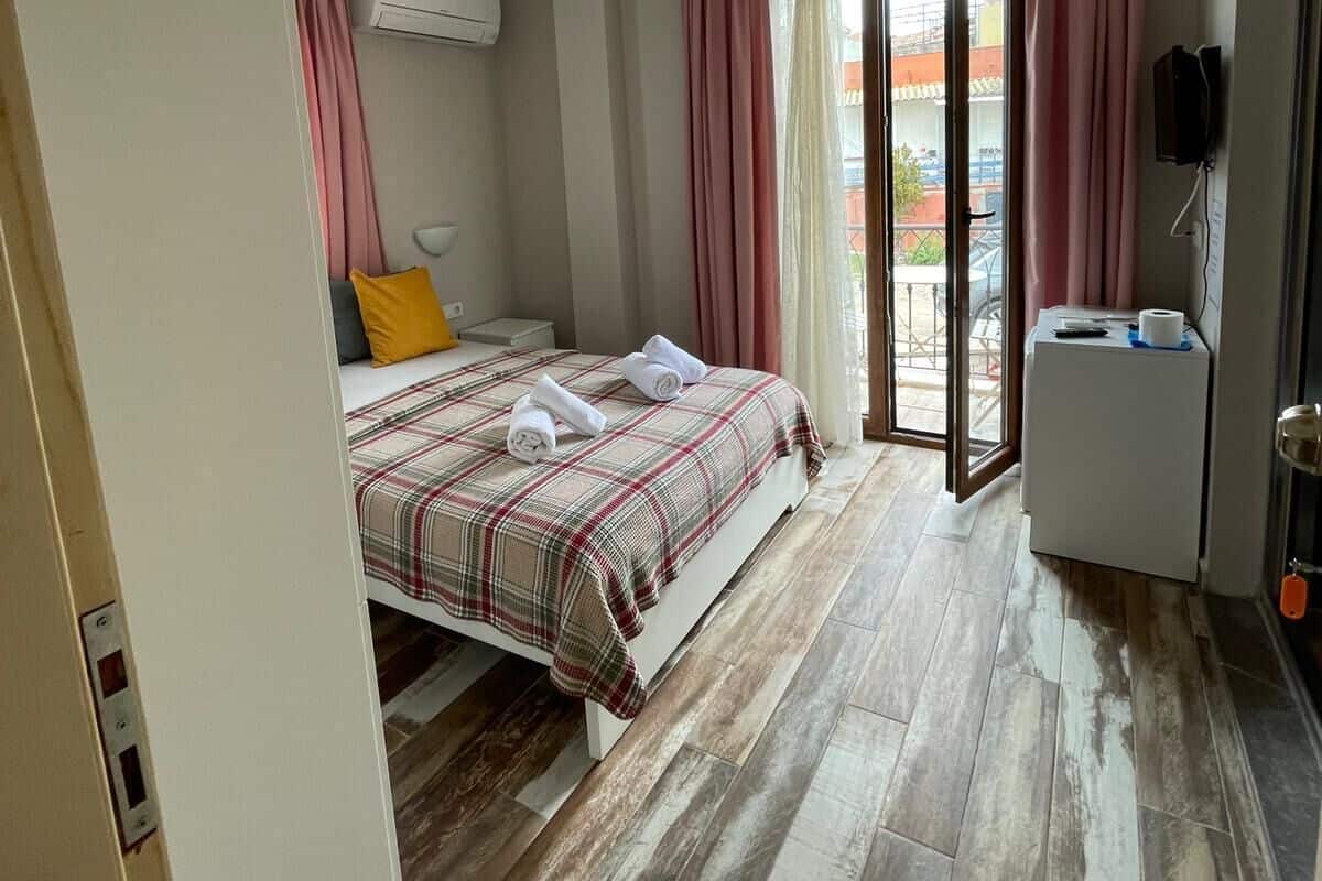A hotel room with a bed, a small table with a remote control, an air conditioning unit, and a sliding glass door leading to an outdoor area.