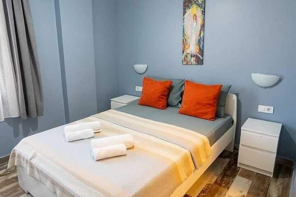 A bedroom with a bed, two nightstands, an air conditioner, a painting, and a window with curtains.