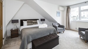 A bedroom with a sloped ceiling, a bed with a grey bedspread, two bedside tables with lamps, a grey armchair, and a window with curtains. - The Porchester Gardens - Modern & Bright 4bdr With Garden and Parking (London)