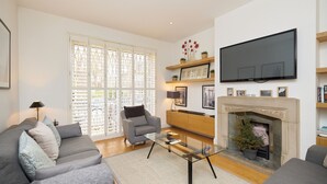 A modern living room with a sofa, armchair, glass coffee table, fireplace, television, and decorative items. - The Porchester Gardens - Modern & Bright 4bdr With Garden and Parking (London)
