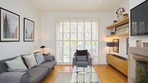 A living room with a grey sofa, a glass coffee table, and a fireplace. There are framed pictures on the wall and a window with a white lattice. - The Porchester Gardens - Modern & Bright 4bdr With Garden and Parking (London)