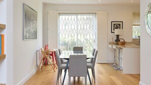 A modern kitchen with a dining area, featuring a large window with a metal grid, a painting on the wall, and a small bar area. - The Porchester Gardens - Modern & Bright 4bdr With Garden and Parking (London)