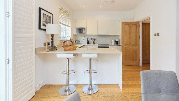 A modern kitchen with white cabinets, a central island with bar stools, a microwave, and a wooden door. - The Porchester Gardens - Modern & Bright 4bdr With Garden and Parking (London)