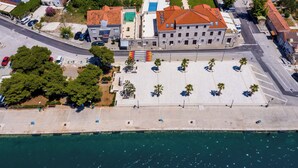 An aerial view of a coastal area with a large parking lot, a building with a red roof, and a waterfront. - Villa Ro-ma (Dubrovacko Primorje)