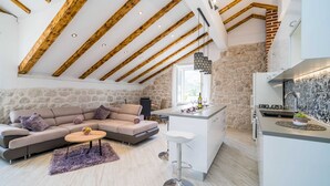 A modern living room with a sofa, a coffee table, and a kitchen area with a stone wall and wooden beams. - Villa Ro-ma (Dubrovacko Primorje)