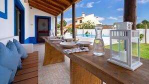 An outdoor dining area with a wooden table set for two, a white lantern, and a view of a swimming pool and buildings in the background. - Amazing Comporta family beach house (Comporta)