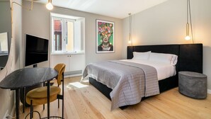 A bedroom with a bed, bedside table, chair, TV, artwork, and a window.