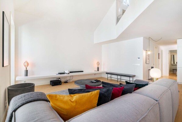 Ein geräumiges Wohnzimmer mit hoher Decke, einem grauen Sofa mit bunten Kissen, einem Couchtisch und einer Bank. Zwei Wandlampen und ein kleiner Tisch mit Lampe sind ebenfalls vorhanden.