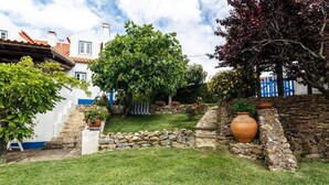 Un jardín bien cuidado con un muro de piedra, un árbol grande y una casa rústica con tejado de tejas.