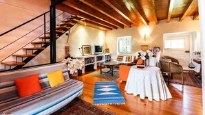 Un salón espacioso con escaleras, suelos de madera, un sofá con cojines, una mesa de comedor y una estantería.
