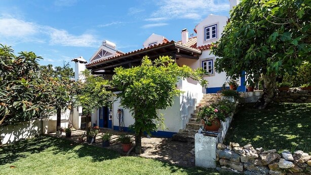 Una casa tradicional con una escalera de piedra que lleva a la entrada, rodeada de un jardín cuidado y un cielo azul despejado.
