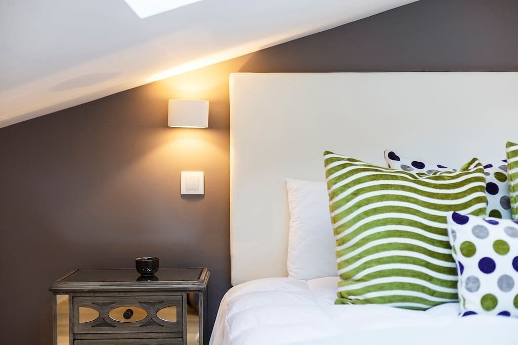 Ein Schlafzimmer mit schrägen Wänden, einem Bett mit weißer Bettwäsche, grün-weißen Streifen-Kissen und einem kleinen Nachttisch mit einer Lampe.
