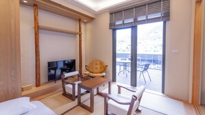 Camera d'albergo con pavimento in legno, tatami, televisore, divano, tavolo e balcone con sedie.