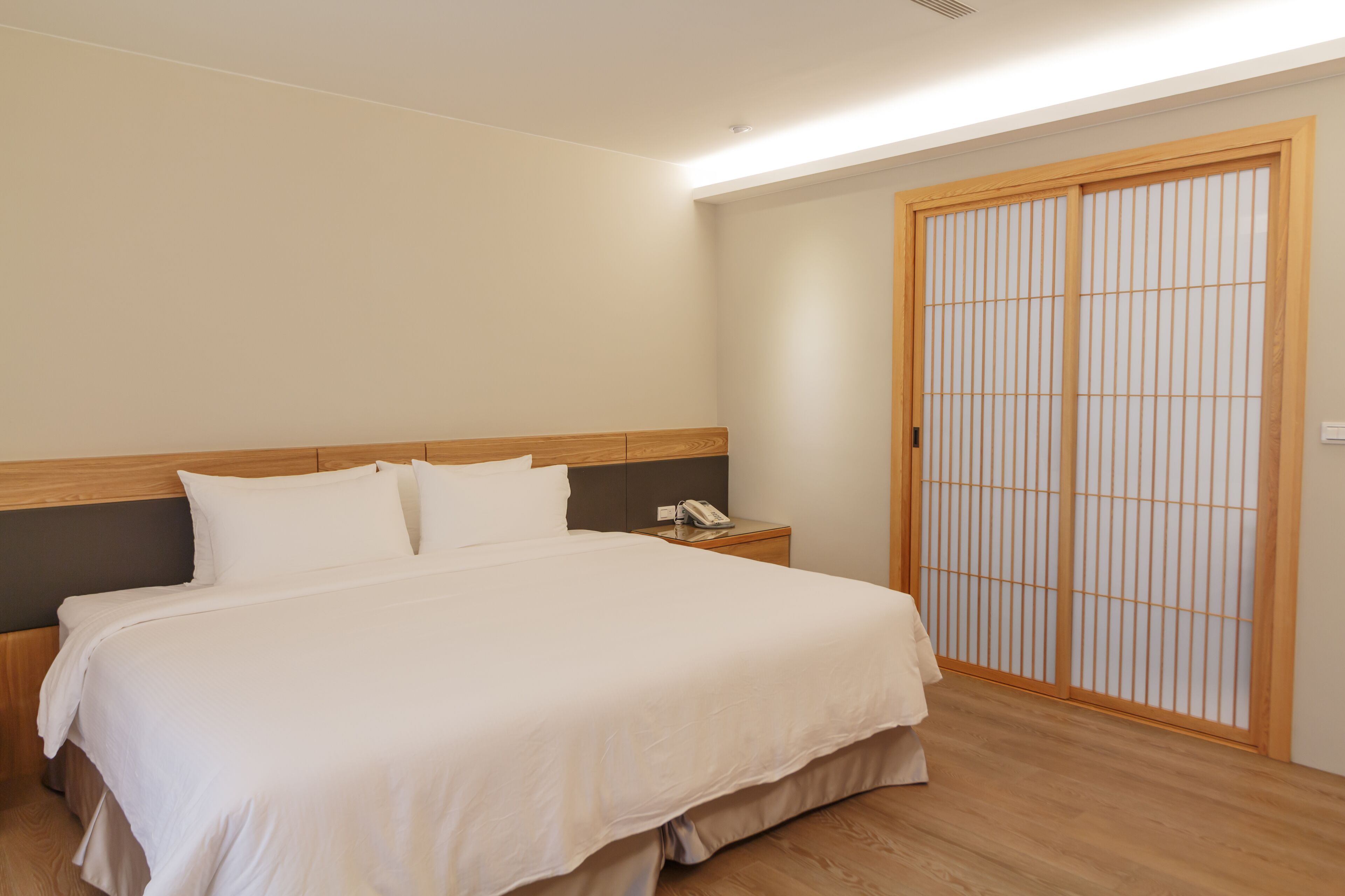 A neatly made bed with a wooden headboard, a bedside table with a telephone, and a sliding shoji screen door.