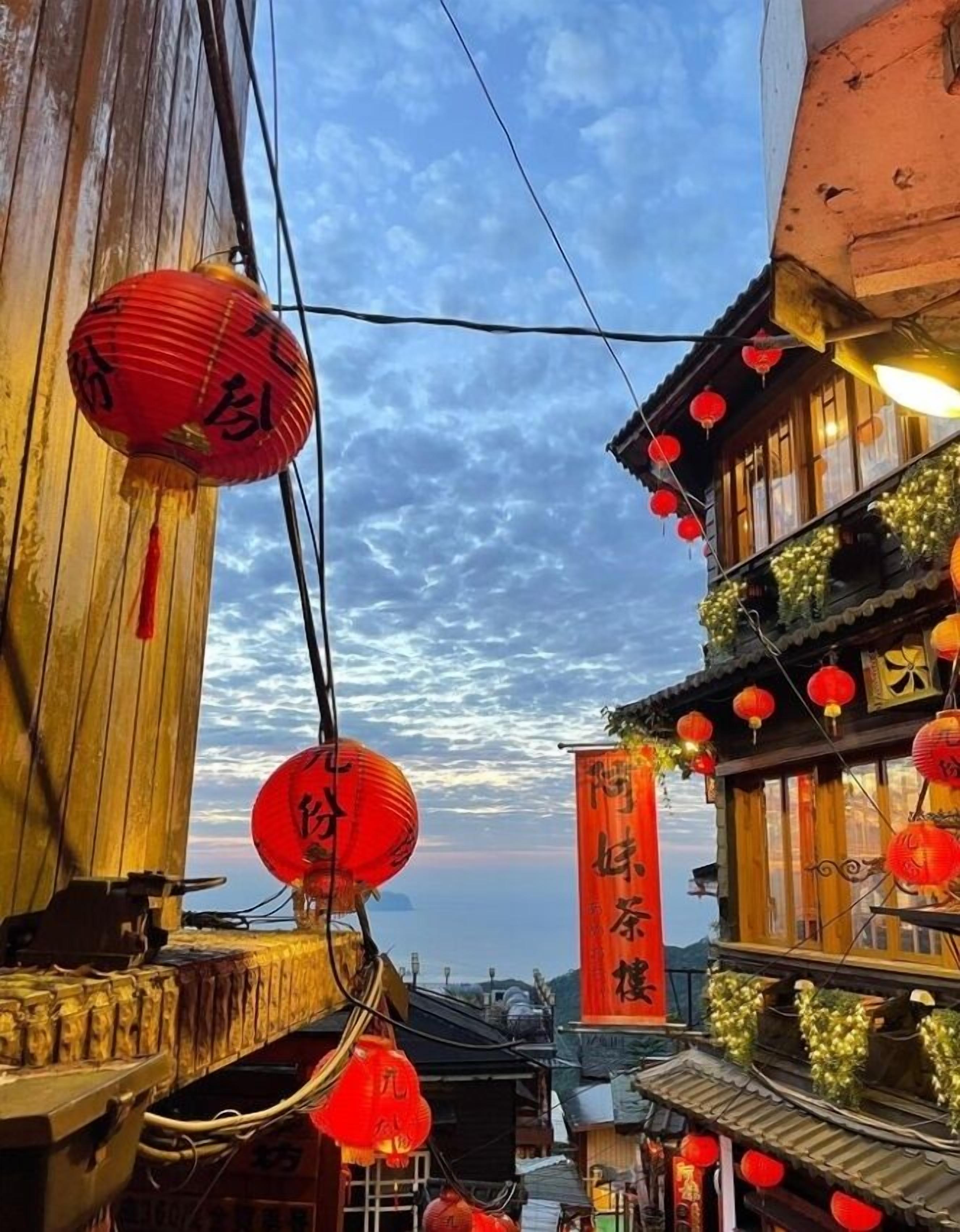A narrow street with traditional wooden buildings, red lanterns, and a clear sky.