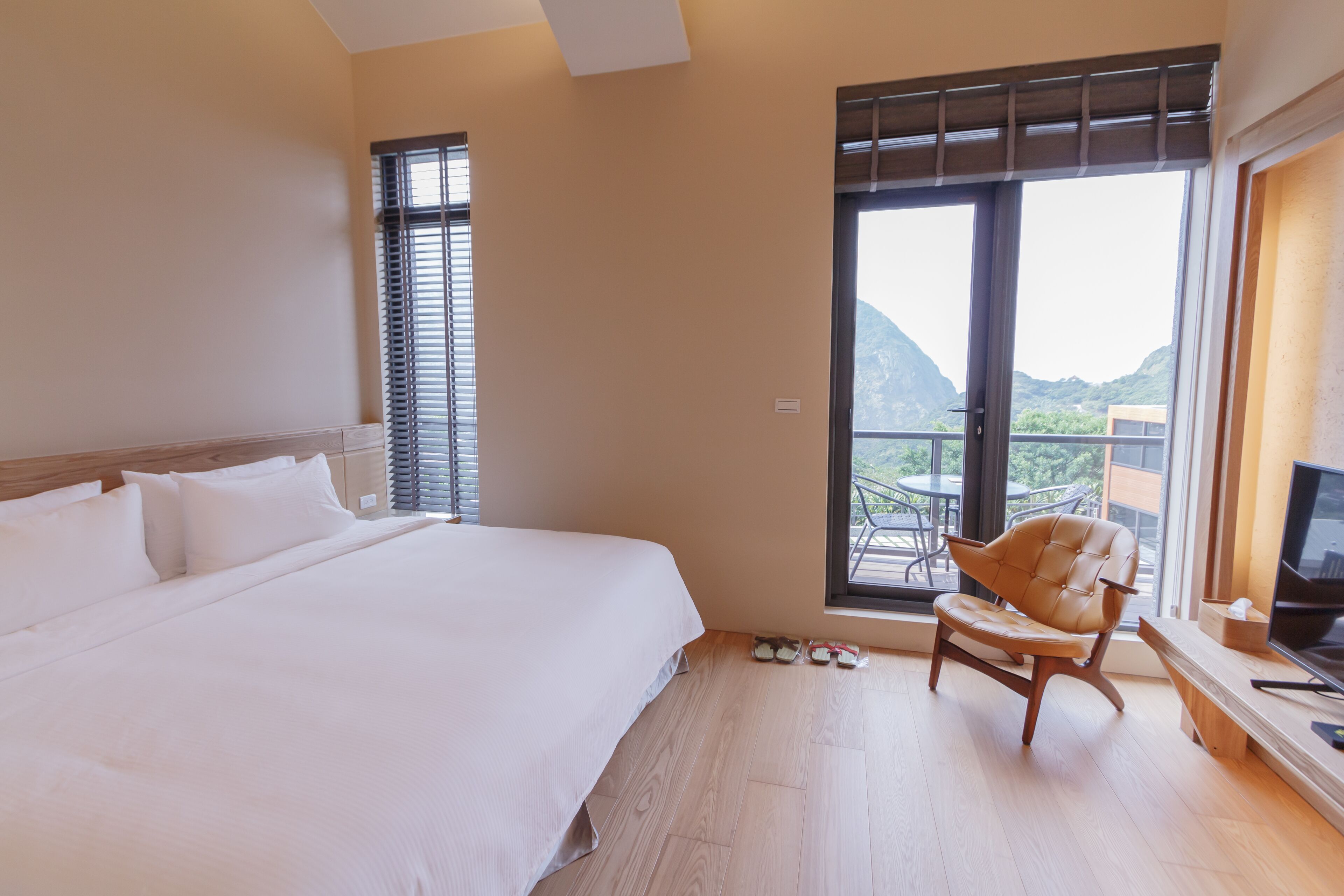 A hotel room with a large bed, a wooden chair, a TV, and a balcony with a mountain view.