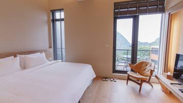 A hotel room with a large bed, a wooden chair, a TV, and a balcony with a mountain view.