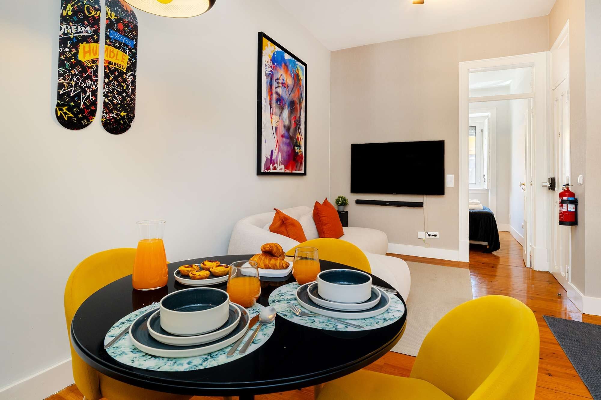 A modern living room with a dining table set for breakfast, a flat-screen TV mounted on the wall, and a fire extinguisher on the wall.