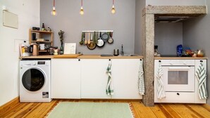 Een praktische keuken met een wasmachine, houten kastjes, hangende pannen en een stenen oven.