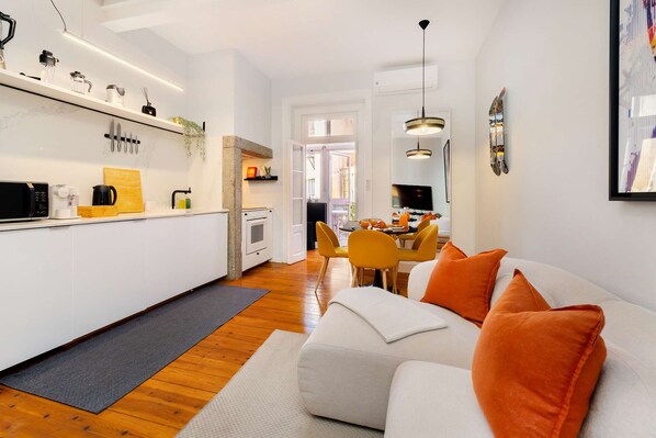A modern kitchen with white cabinets, a wooden countertop, and a dining area with a white sofa and orange cushions.