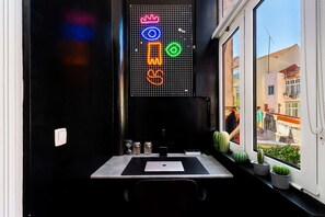 Une pièce avec une fenêtre, un panneau lumineux au néon, une table avec un ordinateur portable et des plantes en pot.