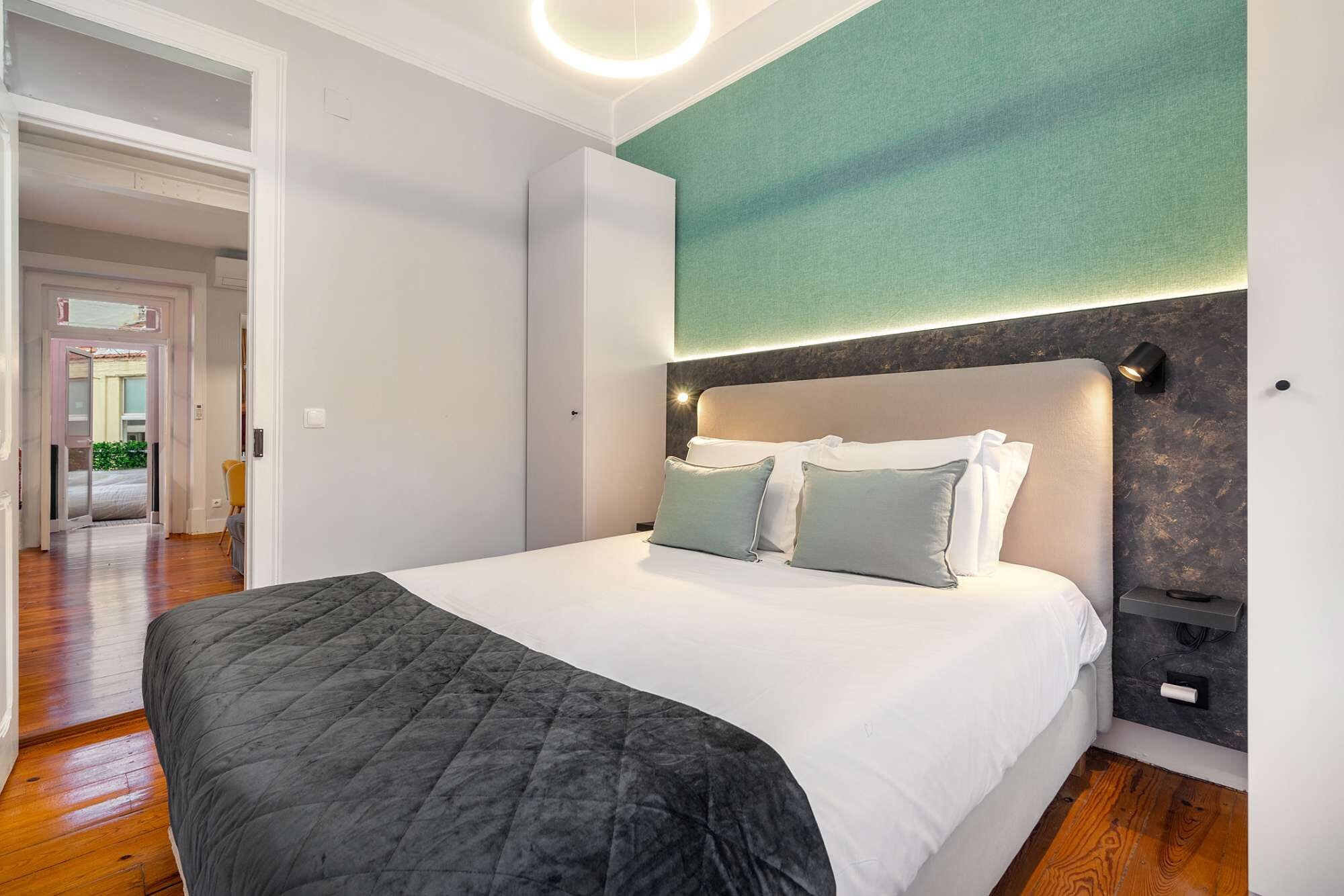 A hotel room with a bed, a bedside table, a wall-mounted light, and a view of the street.