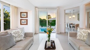 A modern living room with a sofa, a coffee table with a vase of flowers, and a dining area visible through a glass door.