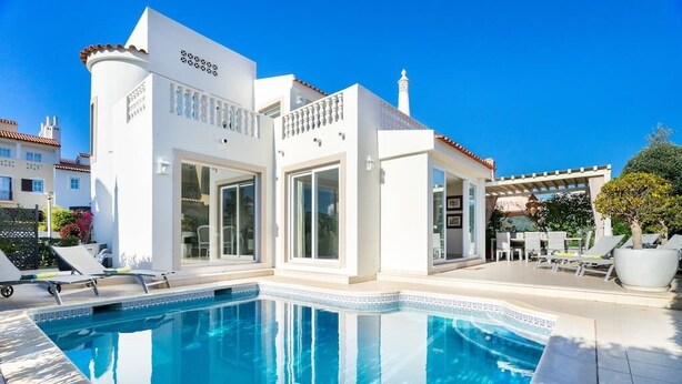 A modern white building with a swimming pool, large windows, and a patio area with lounge chairs.