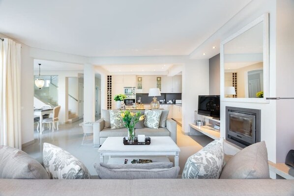 A modern living room with a sofa, coffee table, and a fireplace. There is a kitchen in the background with a microwave and a television. - Prime Hideaway Luxury Villa | Swimming Pool | AC (Quarteira)