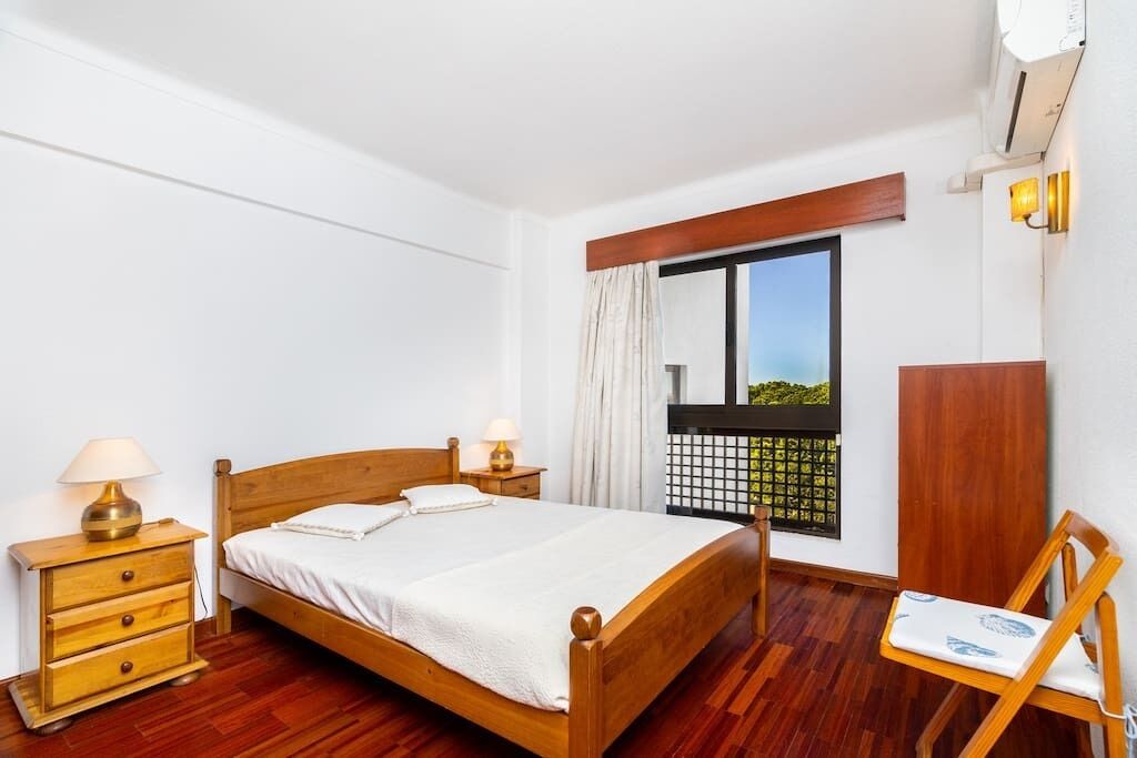 Un dormitorio con una cama de madera, dos mesitas de noche con lámparas, una ventana con vistas y una silla de madera.