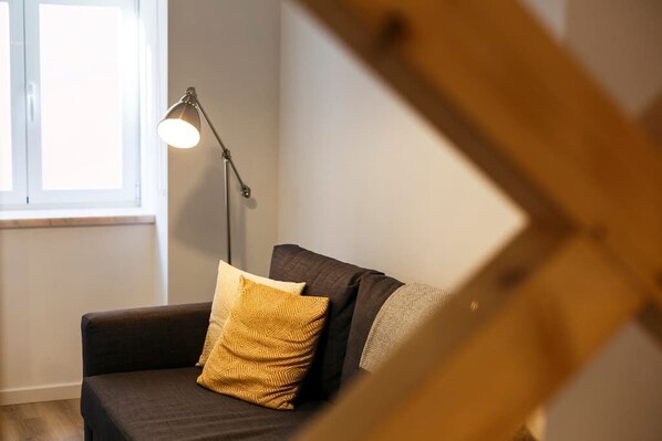 Un sofá de color oscuro con un cojín estampado en una habitación con ventana y lámpara de pie.