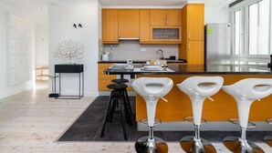 Una cocina moderna con armarios de madera, encimera negra y refrigerador de acero inoxidable. Hay tres taburetes blancos y una mesita negra con un florero.