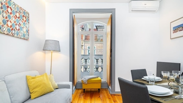 A modern living room with a grey sofa, yellow cushions, a small yellow ottoman, a floor lamp, and a dining area with a table set for two. - Lisbon Prime Aparts - Prata (Lisboa)